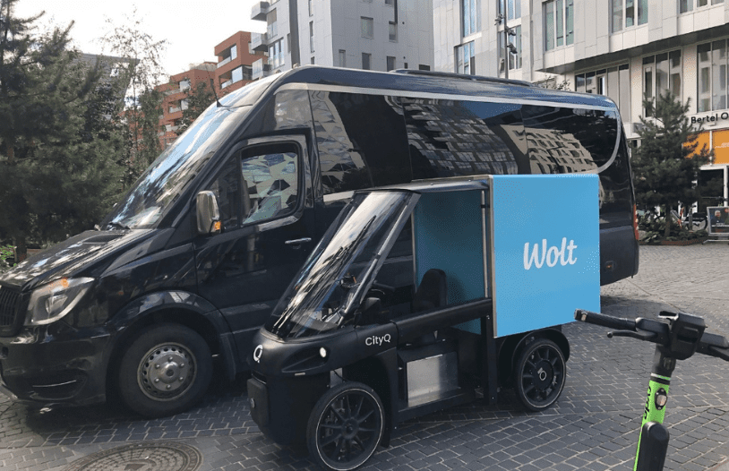 A black CityQ four-wheel cargo e-bike with a blue Wolt delivery box is parked next to a large black van on a city street surrounded by modern buildings.