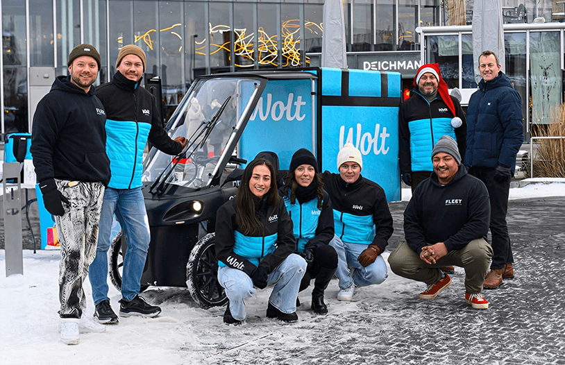 A group of seven Wolt and CityQ employees posing with a branded 4-wheel cargo e-bike in a snowy urban plaza.